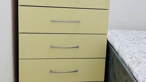 Ein hoher, heller Kommode mit mehreren Schubladen und Griffen, neben einem Bett mit gemustertem Überwurf.