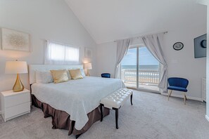 Ein Schlafzimmer mit einem großen Bett, zwei Nachttischlampen, Blick auf den Strand, einer Wanduhr und einem blauen Sessel.