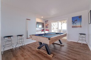 Une table de billard dans une pièce au plancher en bois, avec deux tabourets de bar et un tableau accroché au mur.