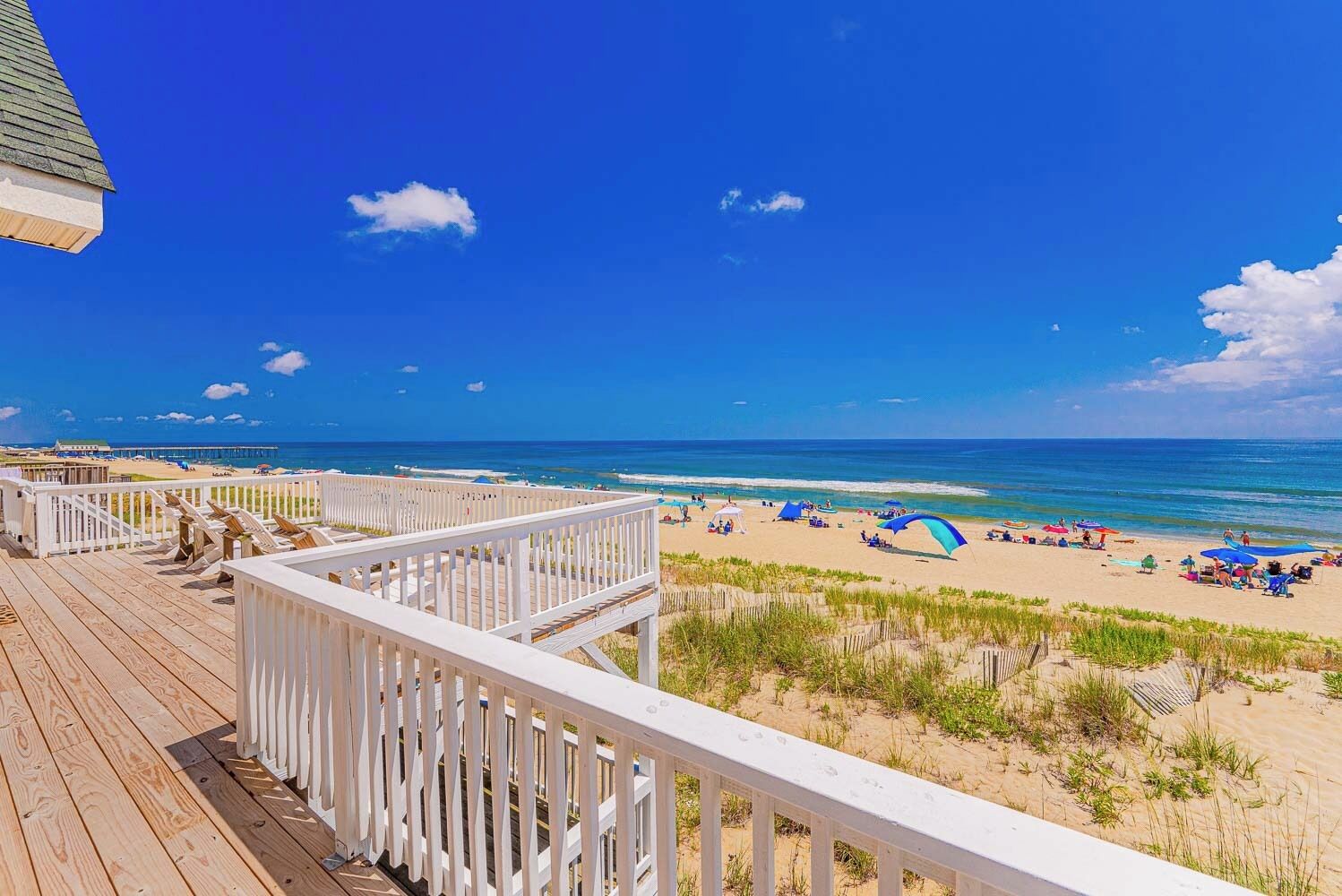 Vue sur la plage depuis une terrasse en bois avec une balustrade blanche, une plage de sable et un ciel bleu dégagé.