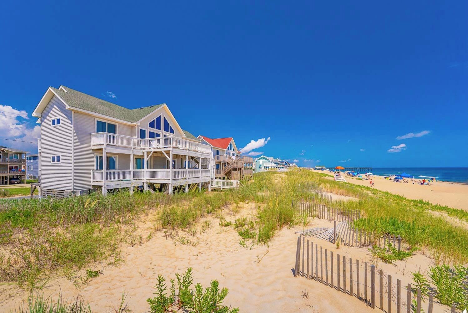 A beachfront property with a large house featuring multiple balconies and a deck, surrounded by sand dunes and beach vegetation.