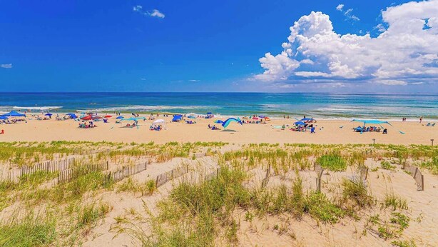 Ein Strand mit Sonnenschirmen und Menschen, Sanddünen und einem klaren Himmel.