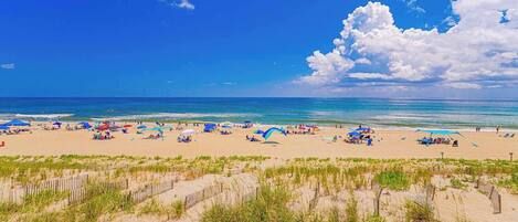 A beach with umbrellas and people, sandy dunes, and a clear sky.