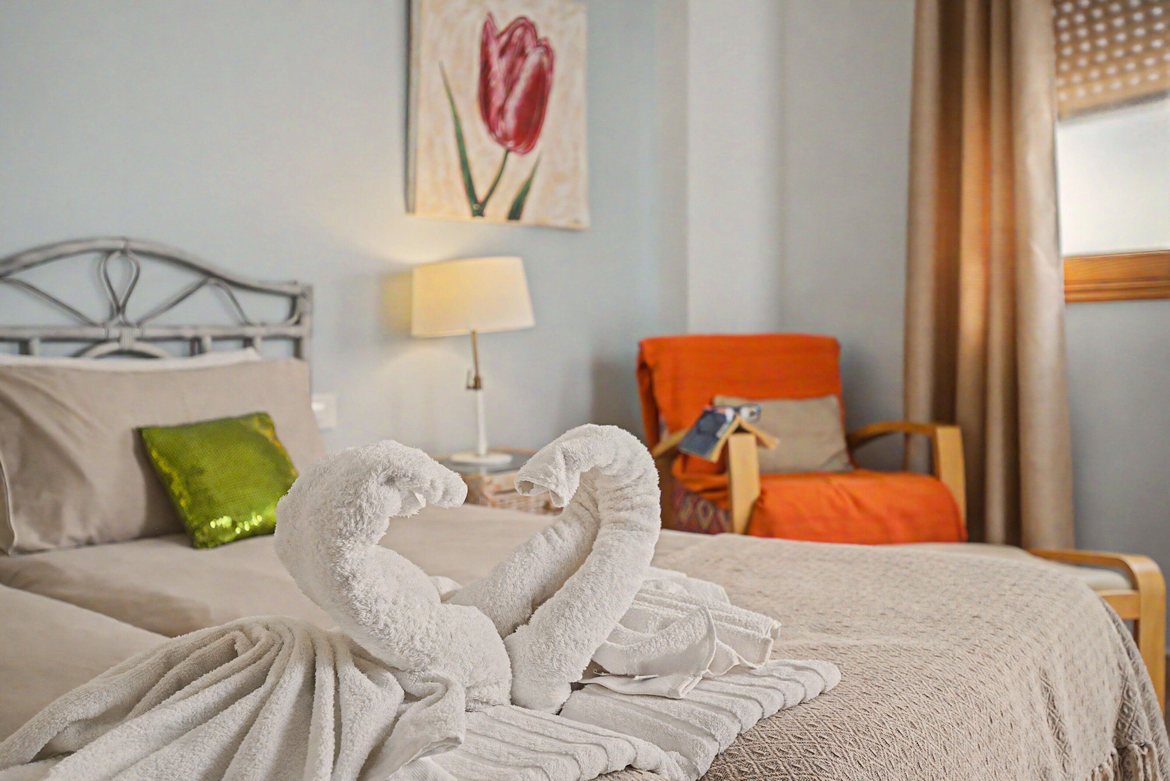 A neatly made bed with white towels arranged in a swan shape, a green pillow, a lamp, and a painting of a tulip on the wall.