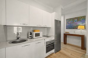 A modern kitchen with white cabinetry, a built-in microwave, a refrigerator, and a wooden side table with a lamp. - Vacation Apartment 'Matias Padron House' with Balcony and Wi-Fi (Las Palmas de Gran Canaria)