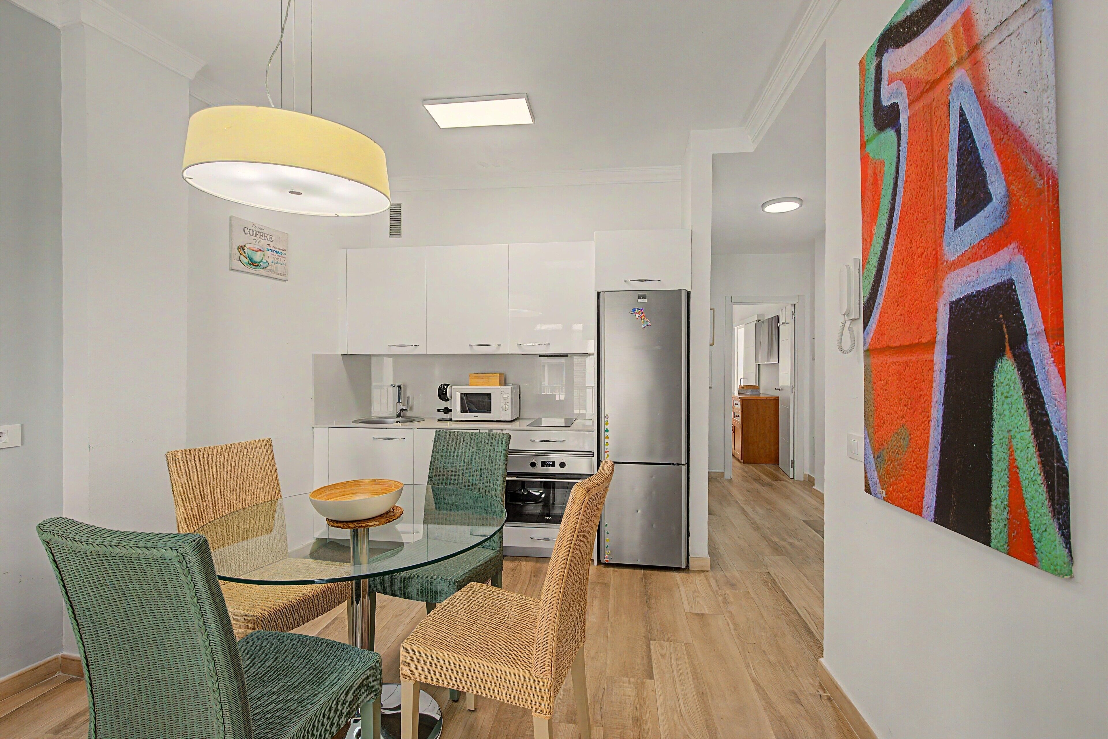 A modern kitchen with white cabinets, a stainless steel refrigerator, and a microwave. There is a dining area with a glass table and wicker chairs.