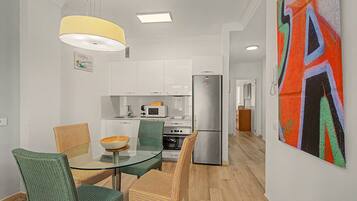 A modern kitchen with white cabinets, a stainless steel refrigerator, and a microwave. There is a dining area with a glass table and wicker chairs.