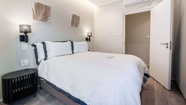 A bedroom with a bed, bedside lamps, a wall-mounted air conditioner, and a door leading to another room. - Jacuzzi, Large Villa, 2 Pools, Camps Bay -Two Views (Cape Town)