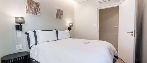 A bedroom with a bed, bedside lamps, a wall-mounted air conditioner, and a door leading to another room.
