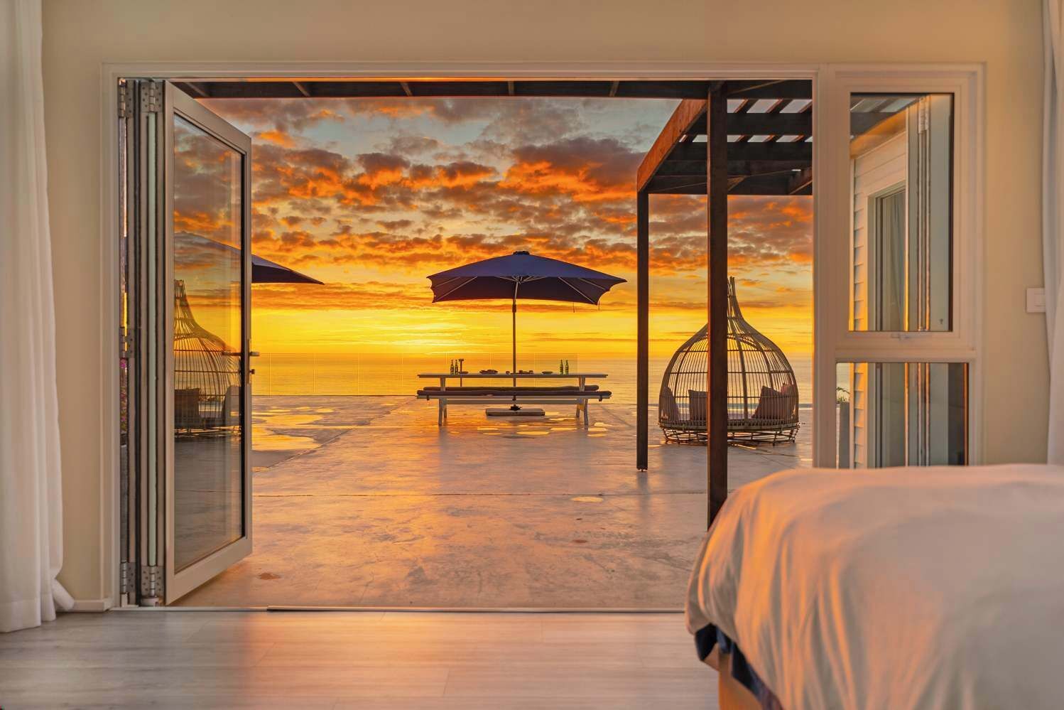 A room with a glass door leading to a beach view, a bed, a table with chairs, and a hanging chair.