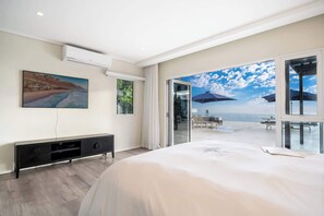 A bedroom with a large bed, a TV, a console table, and a view of the beach through an open door. - Jacuzzi, Large Villa, 2 Pools, Camps Bay -Two Views (Cape Town)
