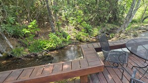 Um deck de madeira com mesa e cadeiras de metal, um churrasqueira e um riacho ao fundo.