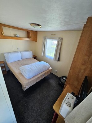 A compact room with a bed, a small table, a heater, and a window with curtains. - Cabin Sleeps 6 With Pets Welcome (Skegness)