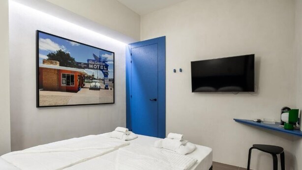Una habitación de hotel con una cama, una puerta azul, un televisor montado en la pared y un cuadro enmarcado de un motel.