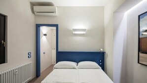 Una habitación de hotel con cama, unidad de aire acondicionado, lámpara y puerta que da a otra habitación.