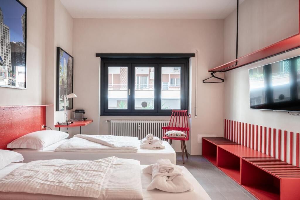 Una camera d'albergo con due letti, una panca rossa, una televisione e una finestra con vista sugli edifici.