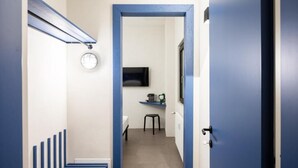 Un pasillo estrecho con un marco de puerta azul, una escalera con barandillas azules, una mesita con una silla y un televisor montado en la pared.