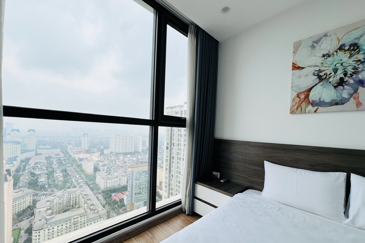 A hotel room with a large window, a bed, a nightstand, and a painting.