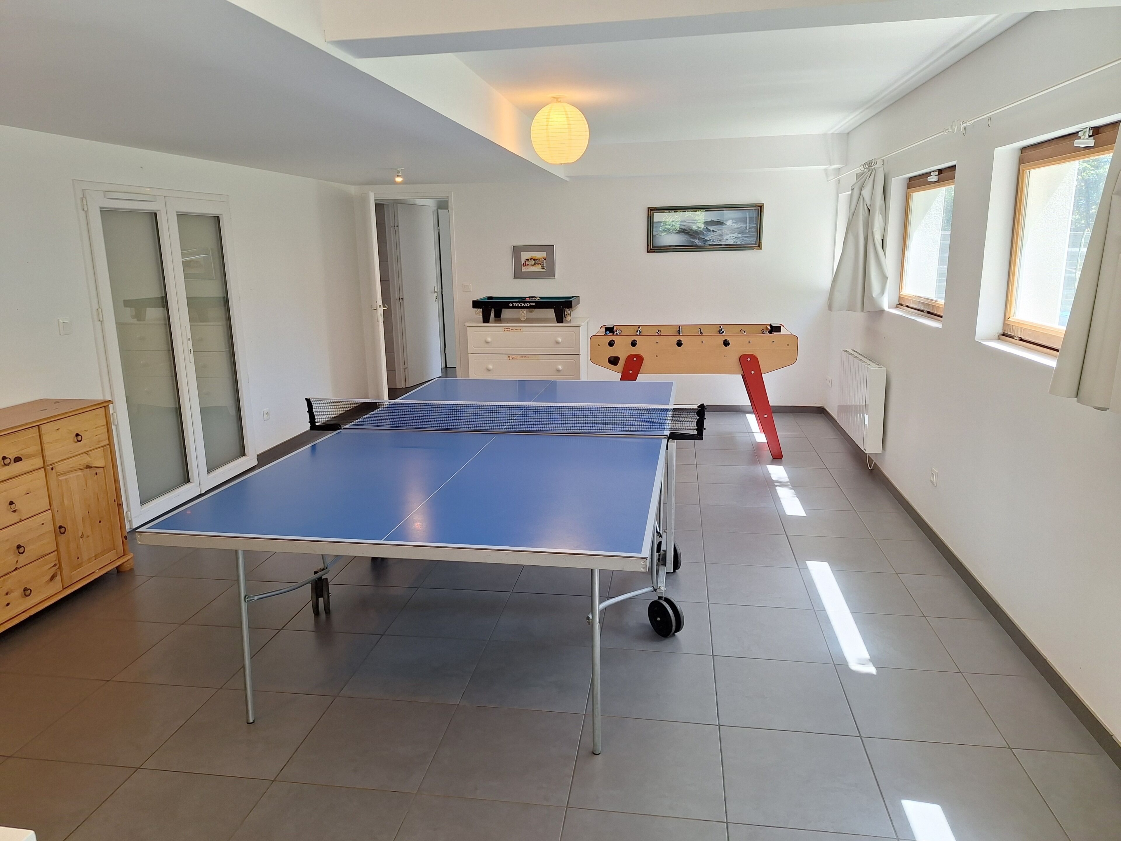Una habitación con mesa de ping pong, un mueble de madera, un radiador y una ventana con cortinas.