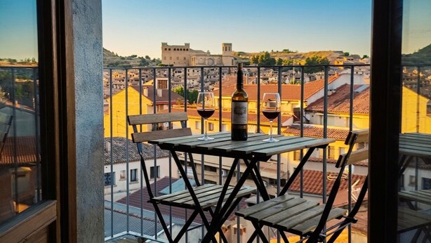 Un balcón con mesa y sillas, con vistas a un paisaje urbano que incluye edificios y una colina.