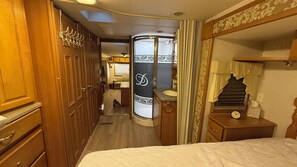 Ein schmaler Flur, der zu einem Badezimmer mit Waschtisch, einem Bett mit Kopfteil und Holzausbau führt.