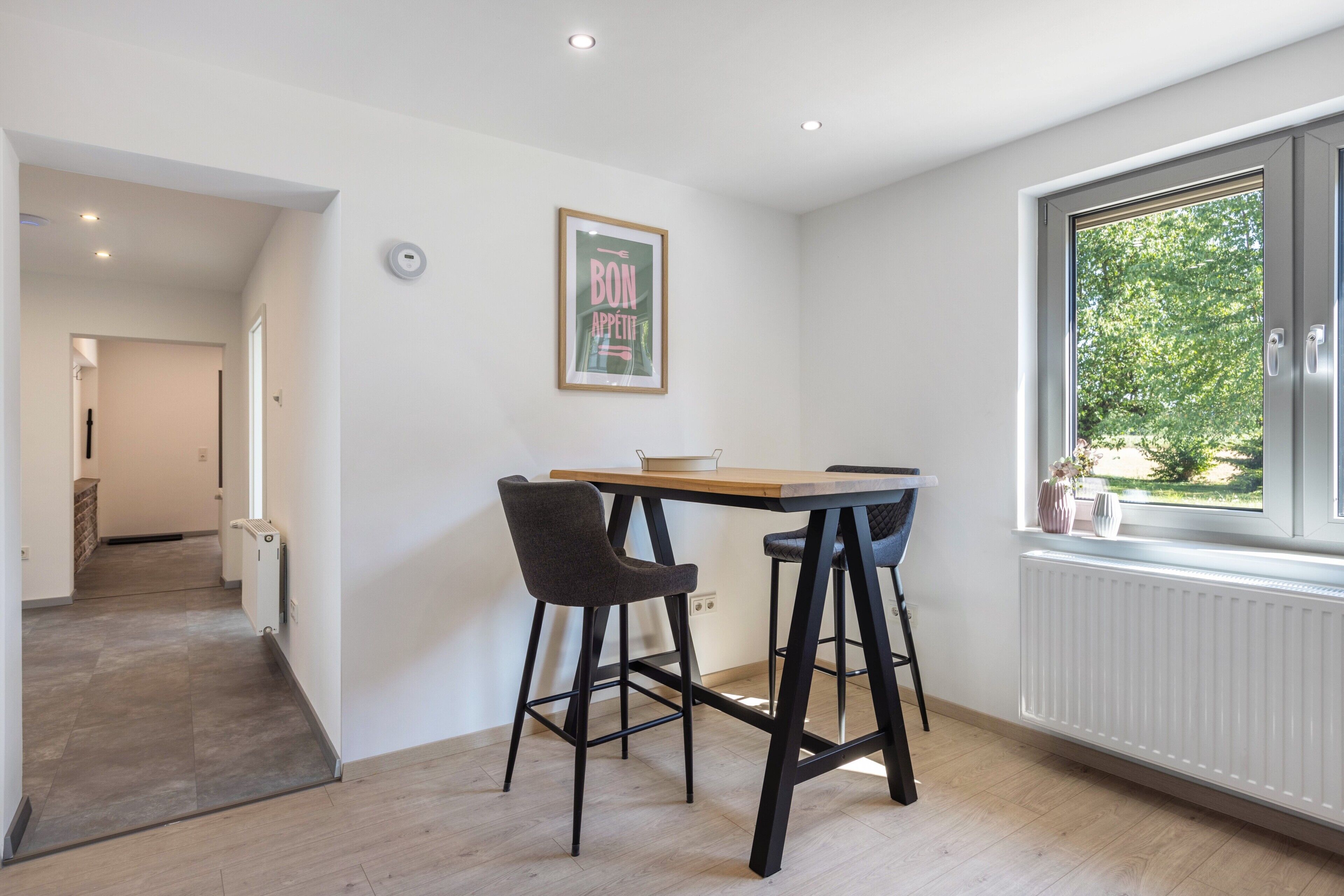 A modern kitchen with a dining area, a framed poster on the wall, and a window with a view of greenery.