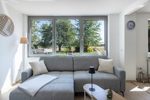 A modern living room with a grey sectional sofa, a small table, and a window with a view of trees and a field. - Apartment "Zur alten Zimmerei" – Vacation in the Werratal (Meinhard)