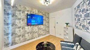 Una sala moderna con televisor de pantalla plana en una pared con textura, una mesa redonda con un jarrón, un sofá con un plaid estampado y una estantería.