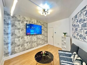 A modern living room with a flat-screen TV mounted on a textured wall, a round coffee table with a vase, a sofa with a patterned throw, and a bookshelf. - South London, Perfectly Placed (London)