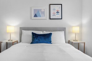 Ein ordentlich bezogenes Bett mit weißer Bettwäsche und einem blauen Kissen, an beiden Seiten zwei Nachttische mit Lampen, an einer schlichten Wand mit zwei gerahmten Kunstwerken.