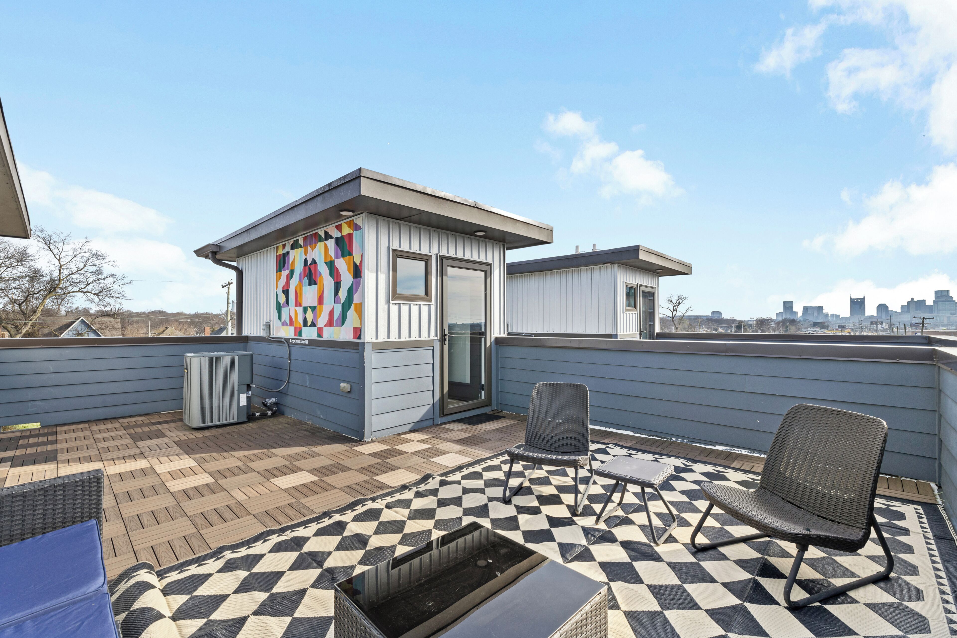 A rooftop patio with a checkered floor, a small building with a colorful wall, and a city skyline in the background.