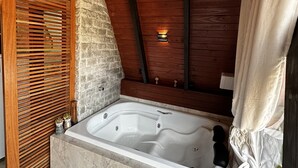 Ein Wellnessbereich mit einer großen Badewanne, Holzbalkendecke, Steinwänden und einer kleinen Wandleuchte.