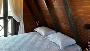 Ein Schlafzimmer mit schr ägem Dach, Holzbalken, einem Bett mit Kissen, einem Fenster mit Vorhängen und einem Nachttisch mit einer Flasche.