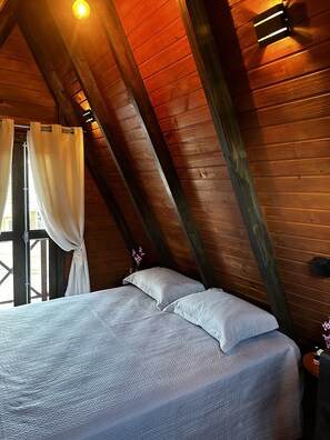 Ein Schlafzimmer mit schrägem Dach, Holzbalken, einem Bett mit Kissen, einem Fenster mit Vorhängen und einem Nachttisch mit einer Flasche.