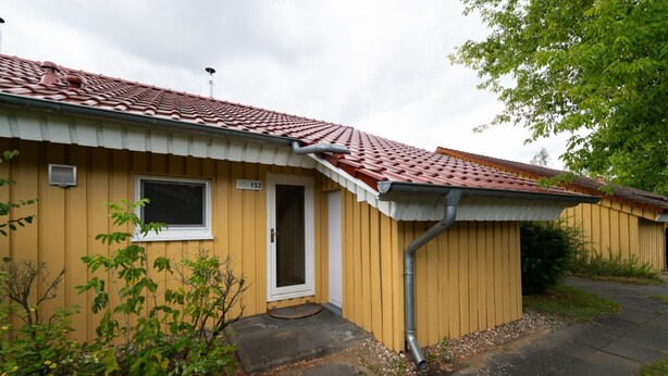 Um edifício amarelo com telhado de telhas vermelhas, uma porta com a inscrição '152' e uma janela.