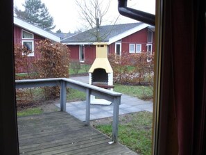 A view of a wooden deck leading to a barbecue area with a chimney, surrounded by a yard with a red building and leaf-covered bushes. - Stilvoll Eingerichteter Doppelbungalow mit Terrasse im Ferienpark Mirow (Mirow)