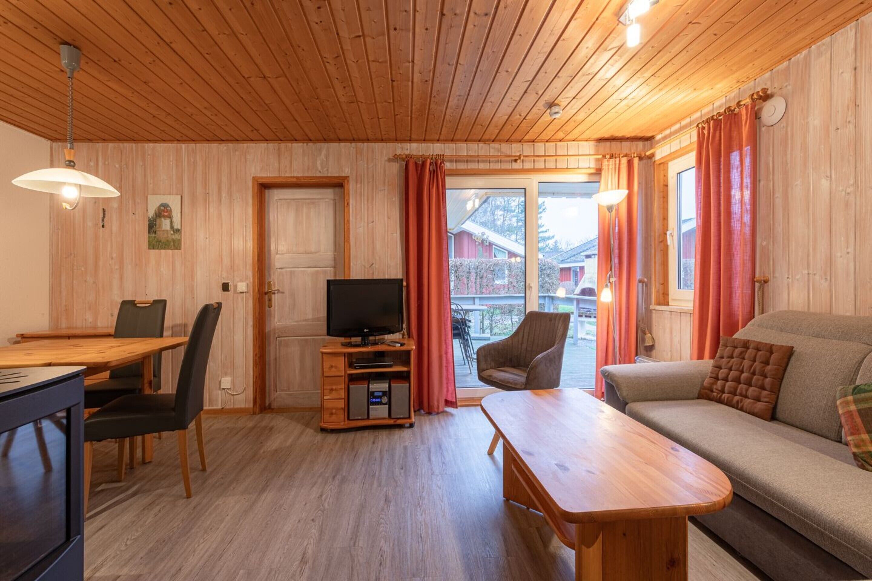 A cozy room with a wooden ceiling, a television on a wooden stand, a sofa, a dining table with chairs, and a view through a glass door.