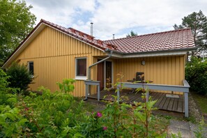 Una casa amarilla con techo de tejas rojas, una terraza de madera y una pequeña mesa al aire libre.