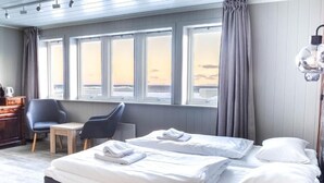 A hotel room with a bed, a chair, a table, and a view of the sea through large windows. - Lofoten Bed & Breakfast (Moskenes)