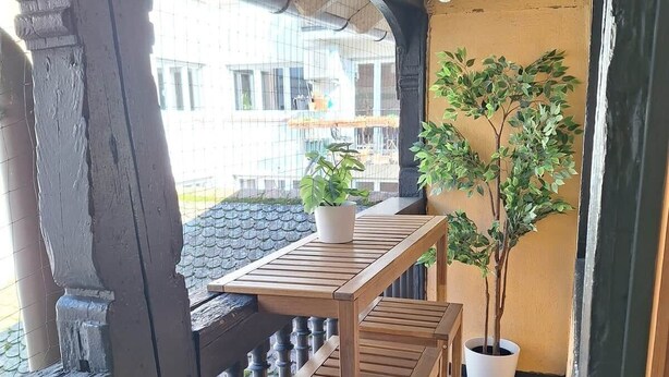 A wooden balcony with a table and chairs, a potted plant, and a view of a building through a wire mesh. - To complete (Strasbourg)