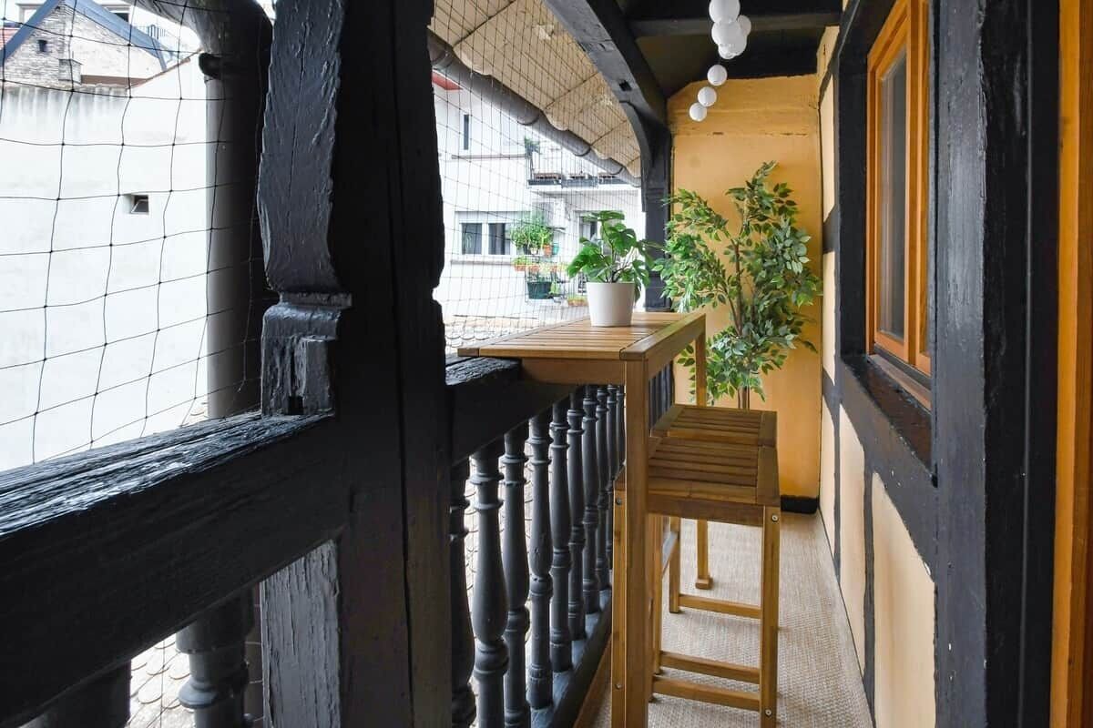 Een smalle gang met een houten bankje, een potplant en uitzicht op de gebouwen buiten.