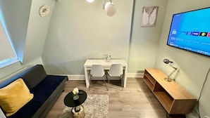 A compact living space with a sofa, a small table, a desk with two chairs, a TV mounted on the wall, and a wooden bench. - Apartment at Albert House, Kennington (London)