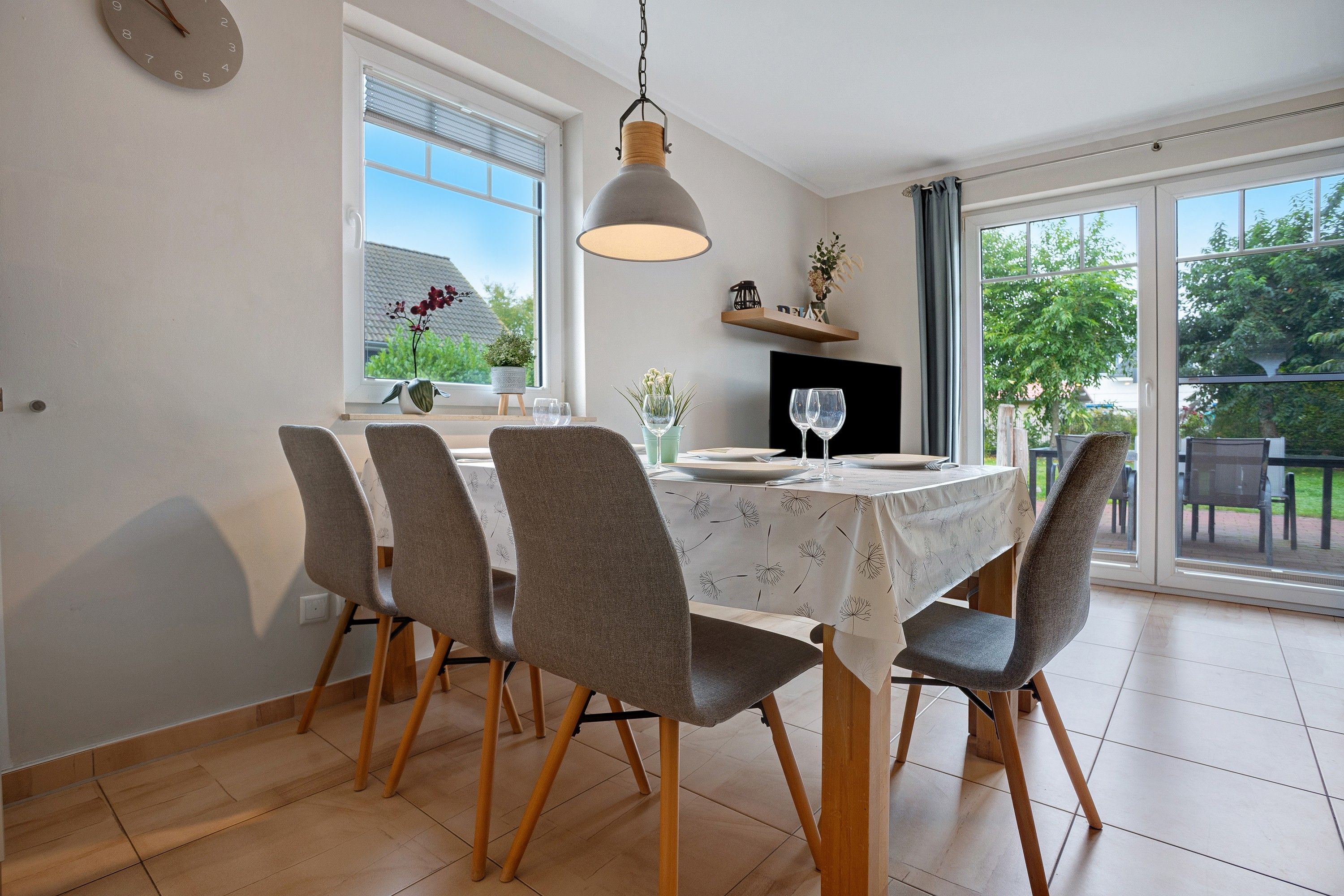 A dining area with a table set for four, chairs, a hanging light fixture, a window with a view, and a shelf with decorative items.