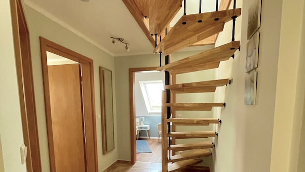 Un pasillo con una escalera de madera que lleva a una zona superior, una puerta a la izquierda y un tragaluz al fondo.
