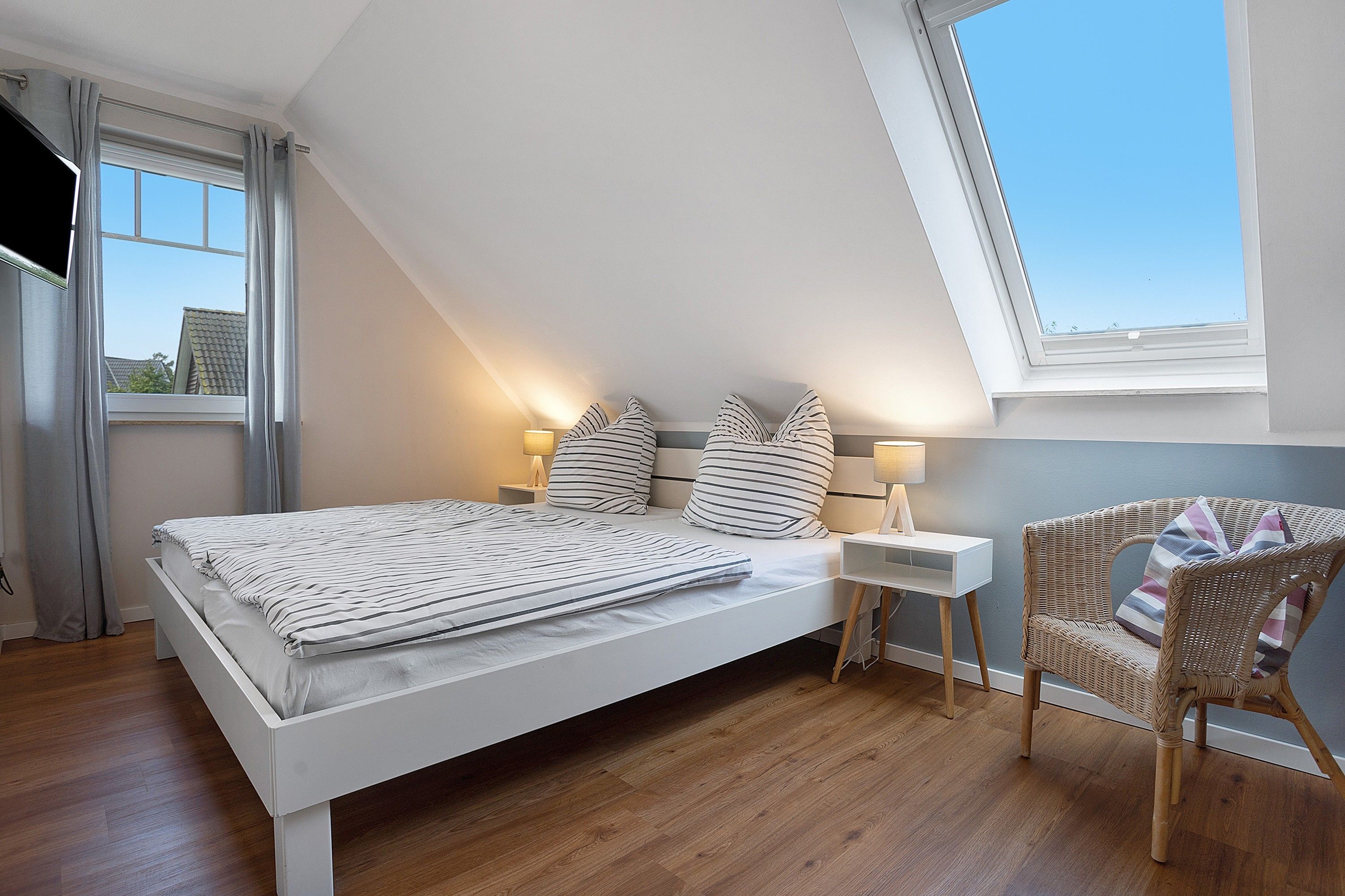 Ein Schlafzimmer mit schrägem Dach, einem Bett mit gestreifter Bettwäsche, zwei Nachttischlampen, einem Korbstuhl und Blick durch ein Fenster.