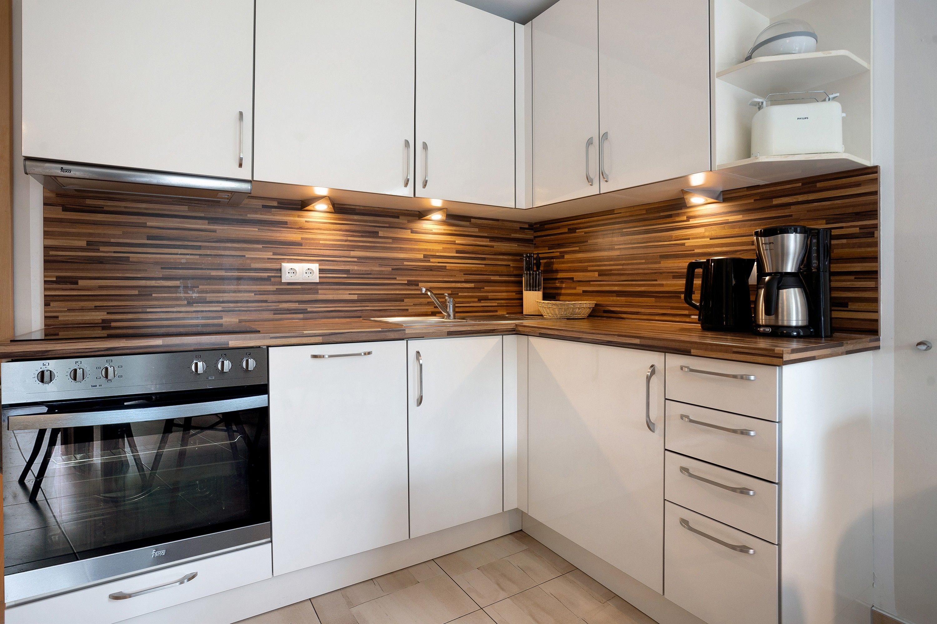 A modern kitchen with white cabinets, a wooden backsplash, a built-in oven, and a coffee maker.