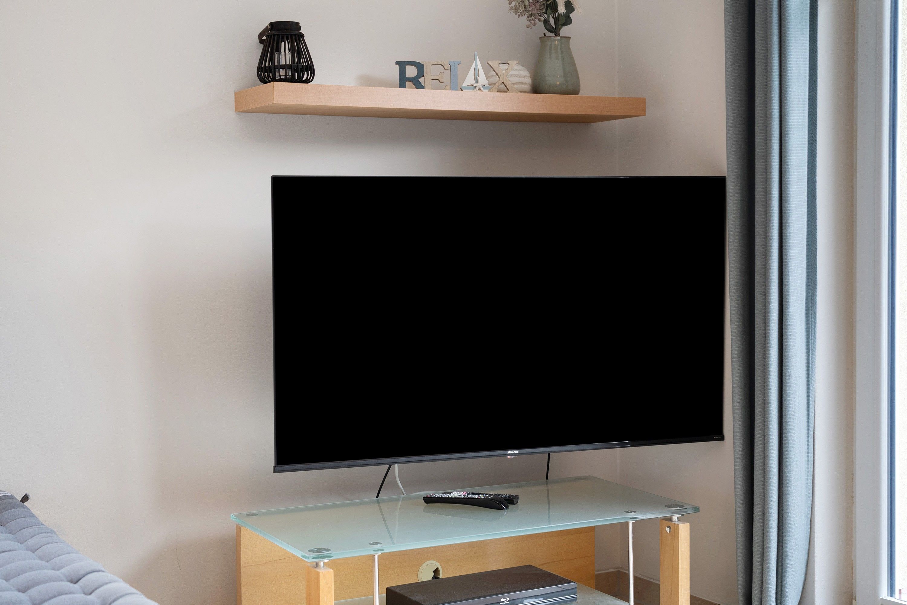 A modern living room with a mounted flat-screen TV, a glass coffee table, and a wooden shelf with decorative items.