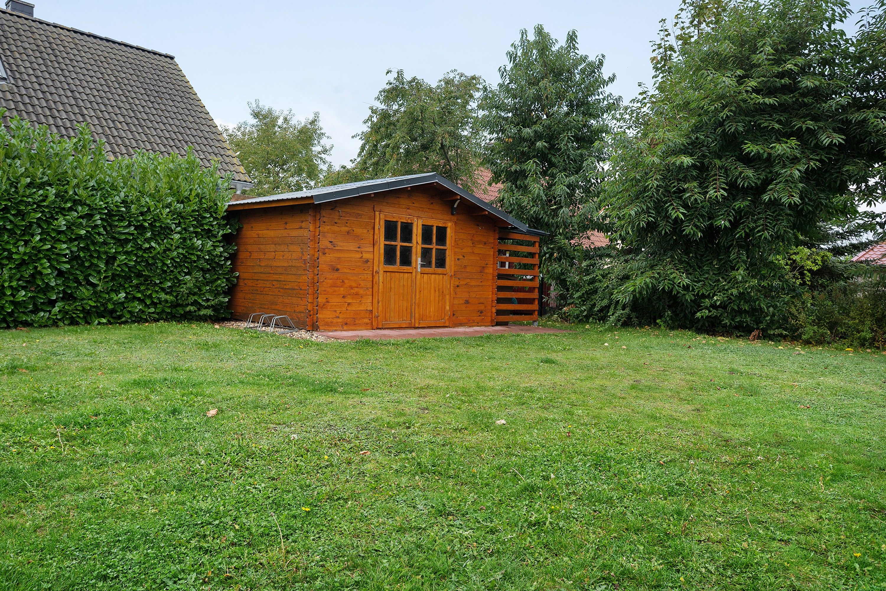 Eine Holzhütte mit Satteldach, einer Tür und einem Fenster, umgeben von einem gepflegten Rasen und dichtem Buschwerk.
