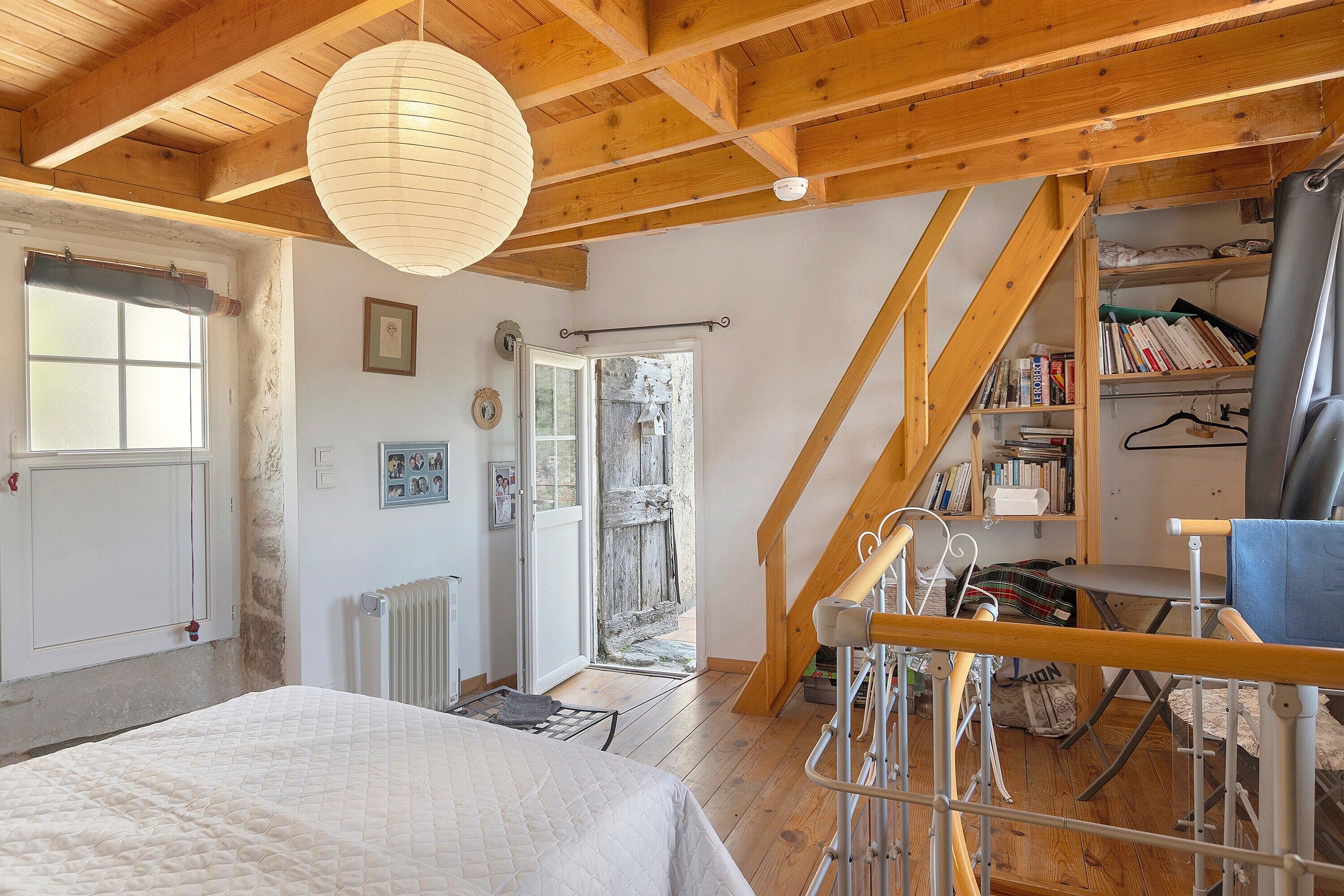 Een slaapkamer met een bed, een radiator, een houten plafond, een raam met jaloezieën en een boekenkast.
