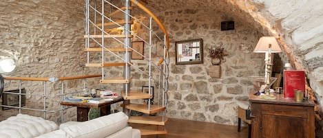 Una habitación con pared de piedra, una escalera de caracol de madera, un sofá, un escritorio con lámpara y una estantería.